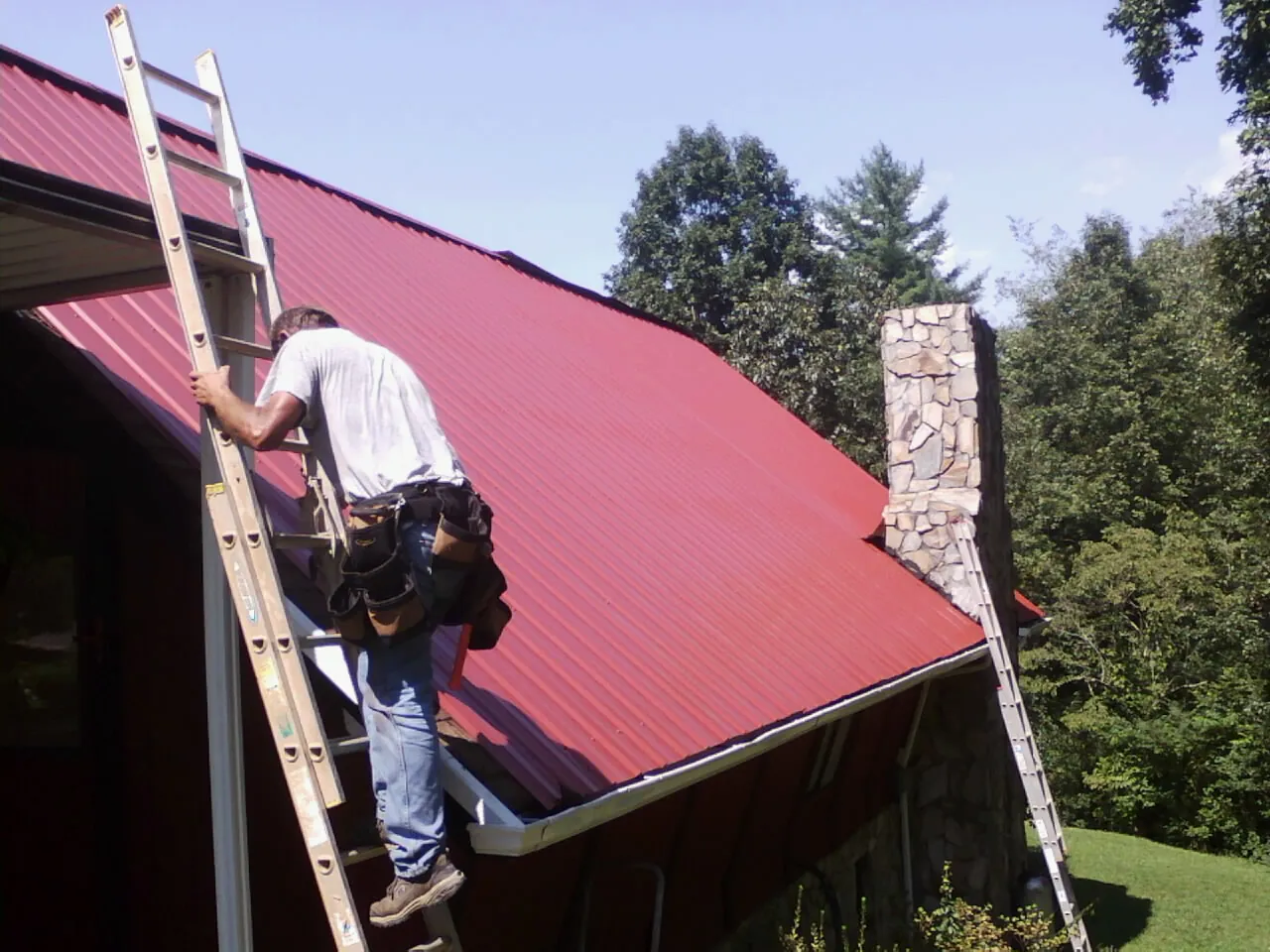 Roofer on a ladder beside a red metal roof and stone chimney — Soffit & Fascia Repair by Roofing Brothers in Ashwood
